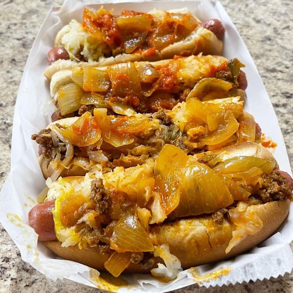 Four hot dogs in buns, topped with chili, sauerkraut, and sautéed onions, in a paper tray.