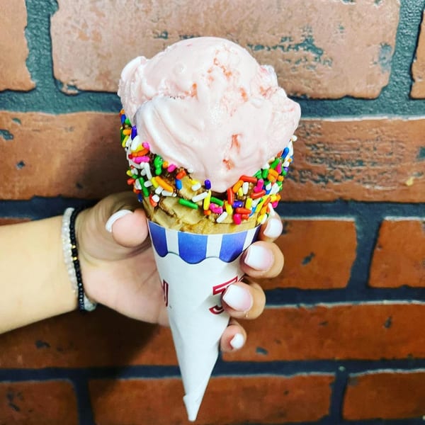 A hand holds a waffle cone with pink ice cream and colorful sprinkles against a brick wall.